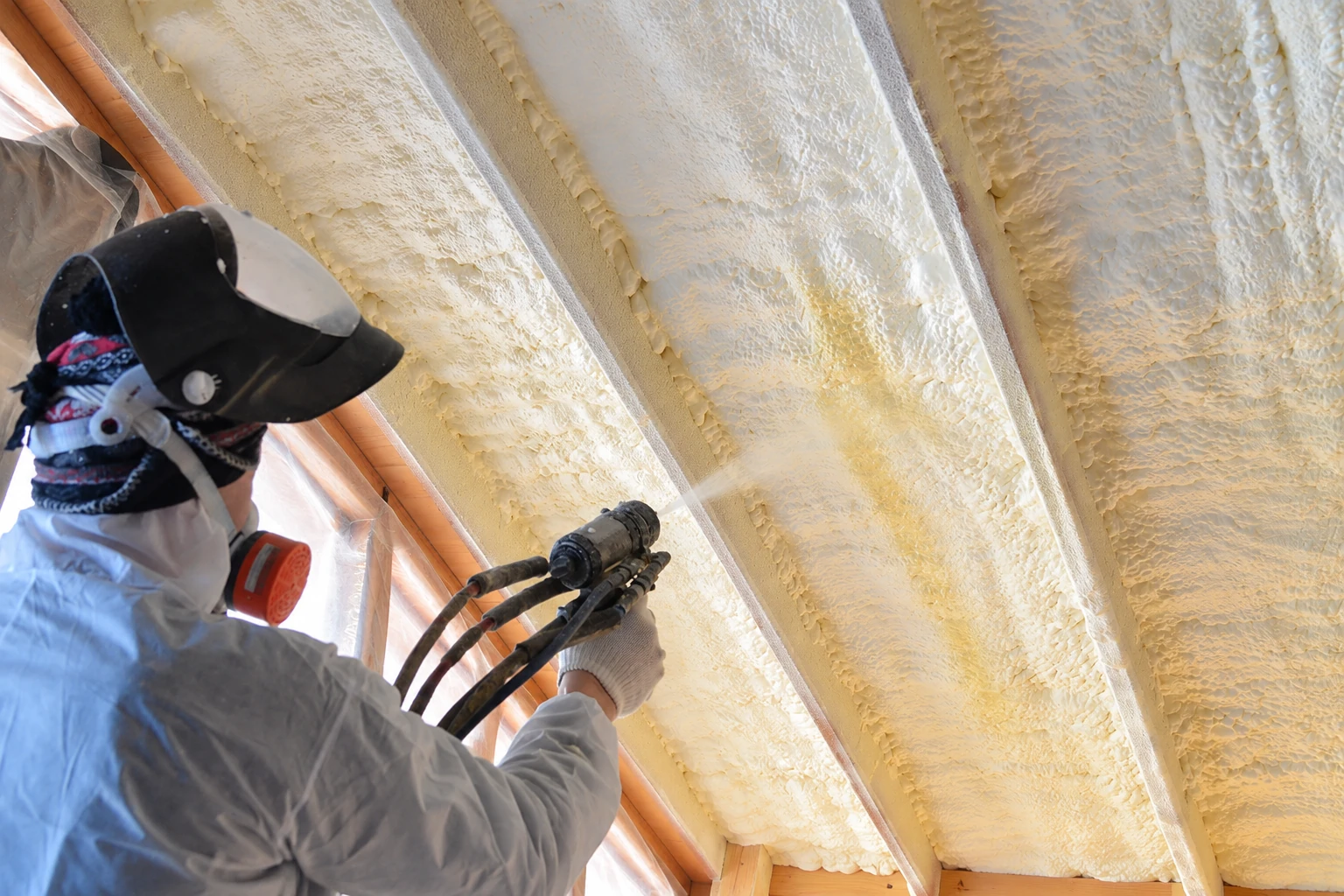 A worker in protective gear sprays foam insulation into the ceiling cavities of a wood-framed roof.
