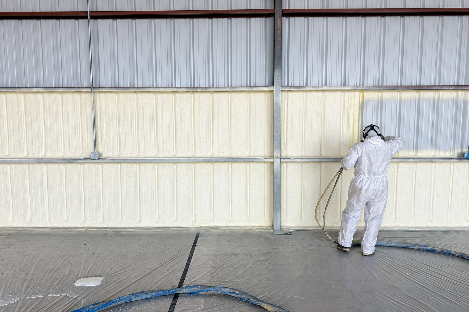 A worker in protective coveralls sprays pale foam insulation onto the wall of a metal commercial building.