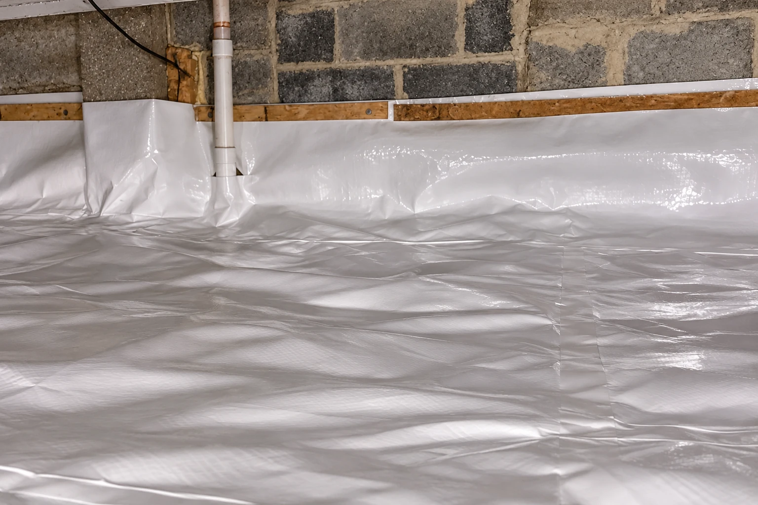 A white vapor barrier covers the crawl space floor and lower block walls around a pipe.