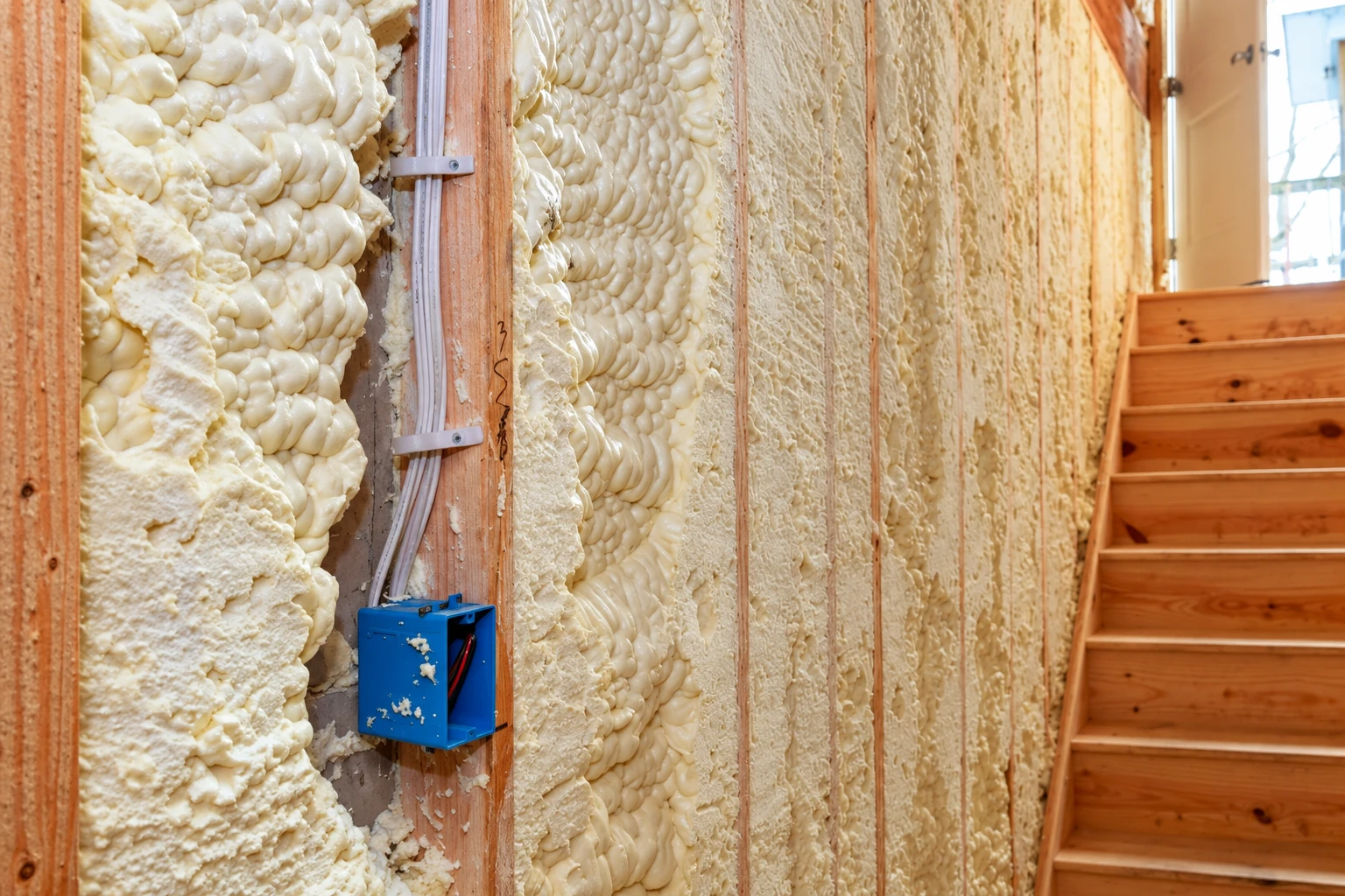 Open-cell foam insulation fills the exposed wall cavities between wooden studs, beside a blue electrical box.