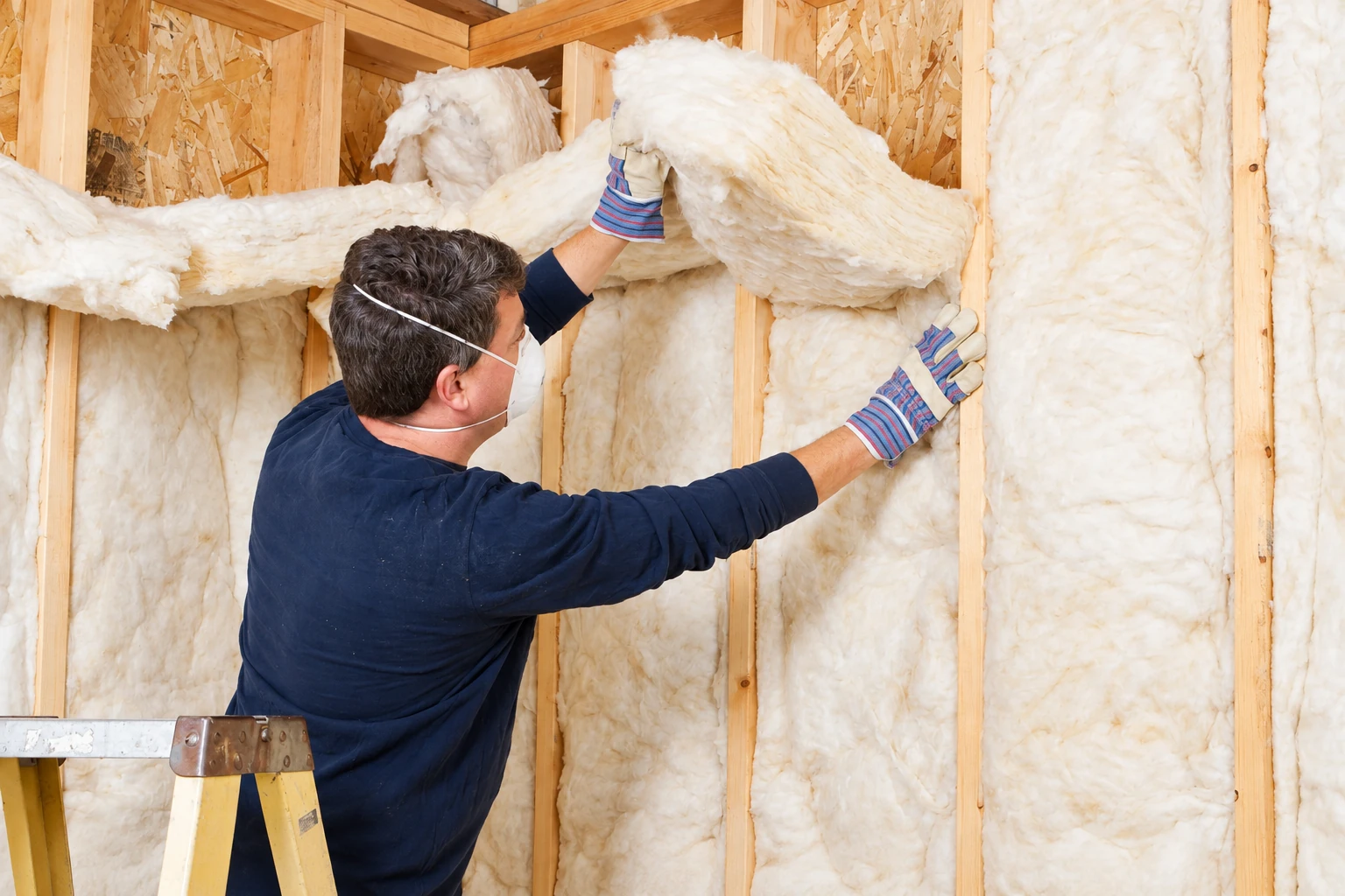 Worker in a mask and gloves fits fluffy white batt insulation between wooden wall studs.